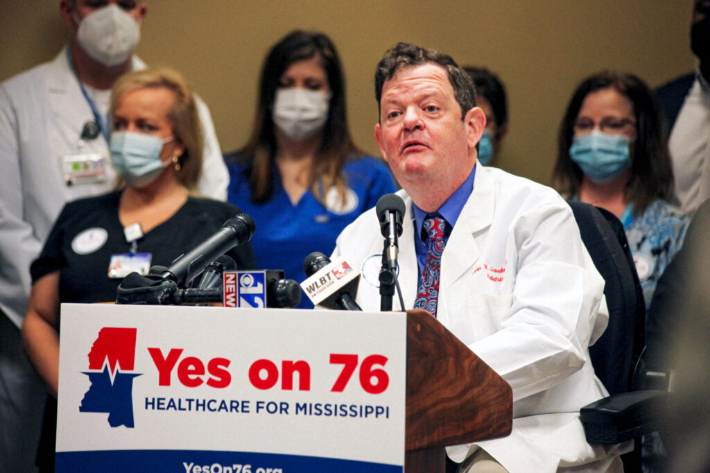 Dr. John Gaudet stands at the podium with nurses and health workers behind him and a YES ON 76 campaign sign in front of him in support of Medicaid expansion
