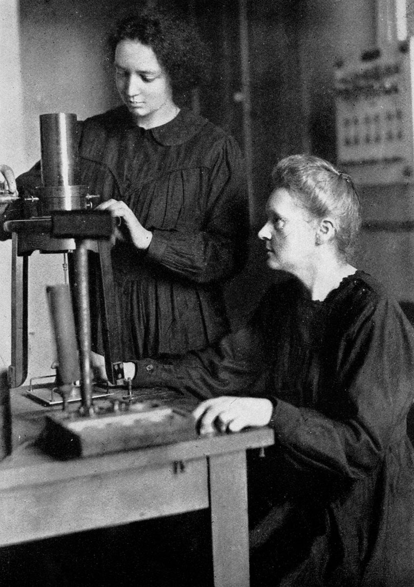 Black and white image of Marie Curie sitting in front of a work table watching as her daughter adjusts an instrument.