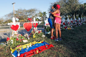Two mourners hug in front of a long row of crosses with flowers resting at the bases