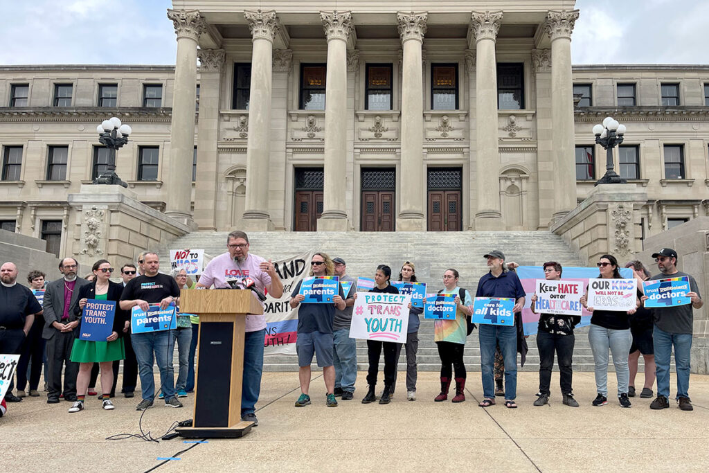 Several people stand in front of the Mississippi Capitol Building holding signs such as "Protect Trans Youth" and "Keep Hate out of Healthcare"