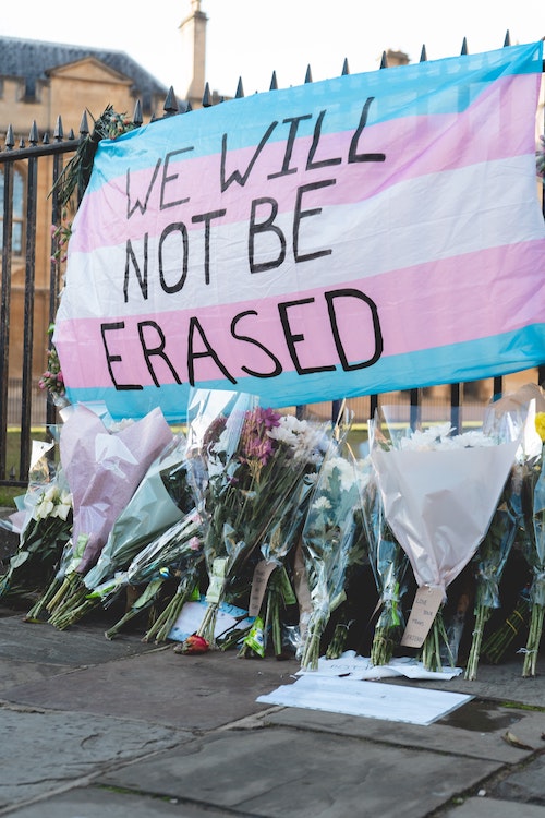 Memorial on fence with trans flag reading, "WE WILL NOT BE ERASED" hanging on a fence with flowers. (trans youth)