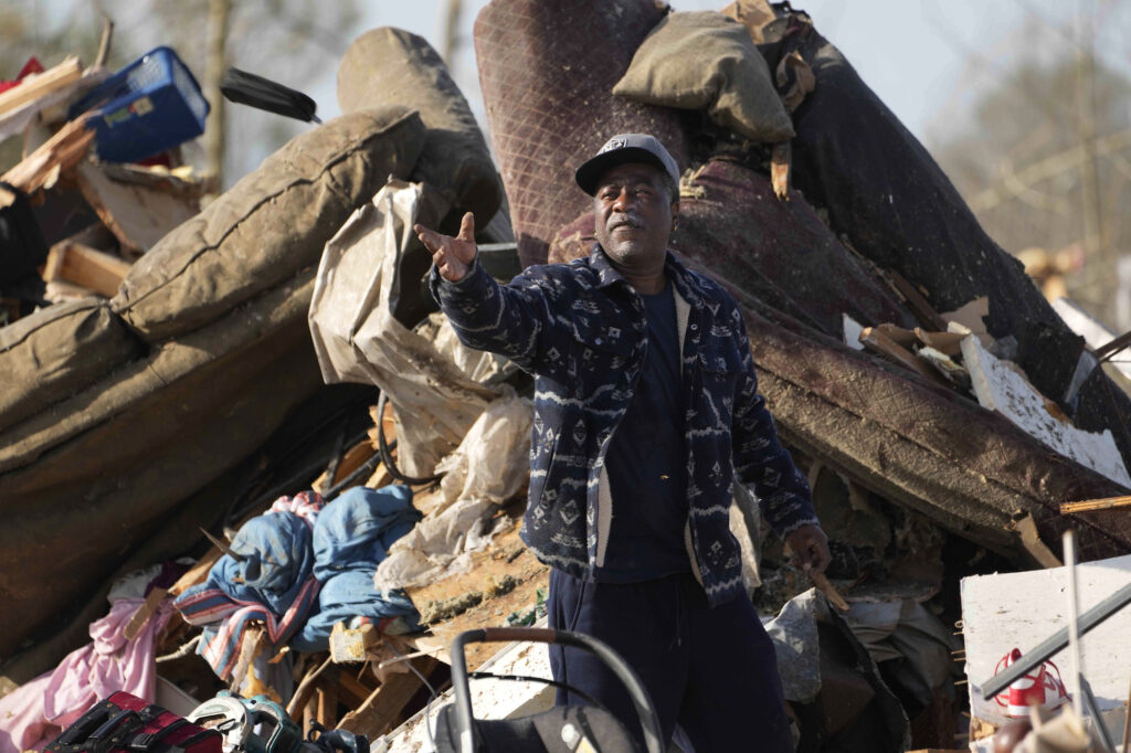 a photo of a man with a gray beard standing amid piles of debris that include sofas and insulation, and holding his hands out, a baseball cap atop his head