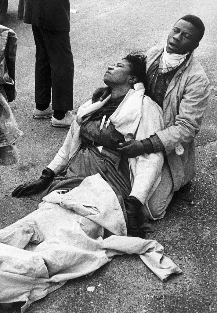 A man crouching on the pavement cradles an injured woman (Black women)