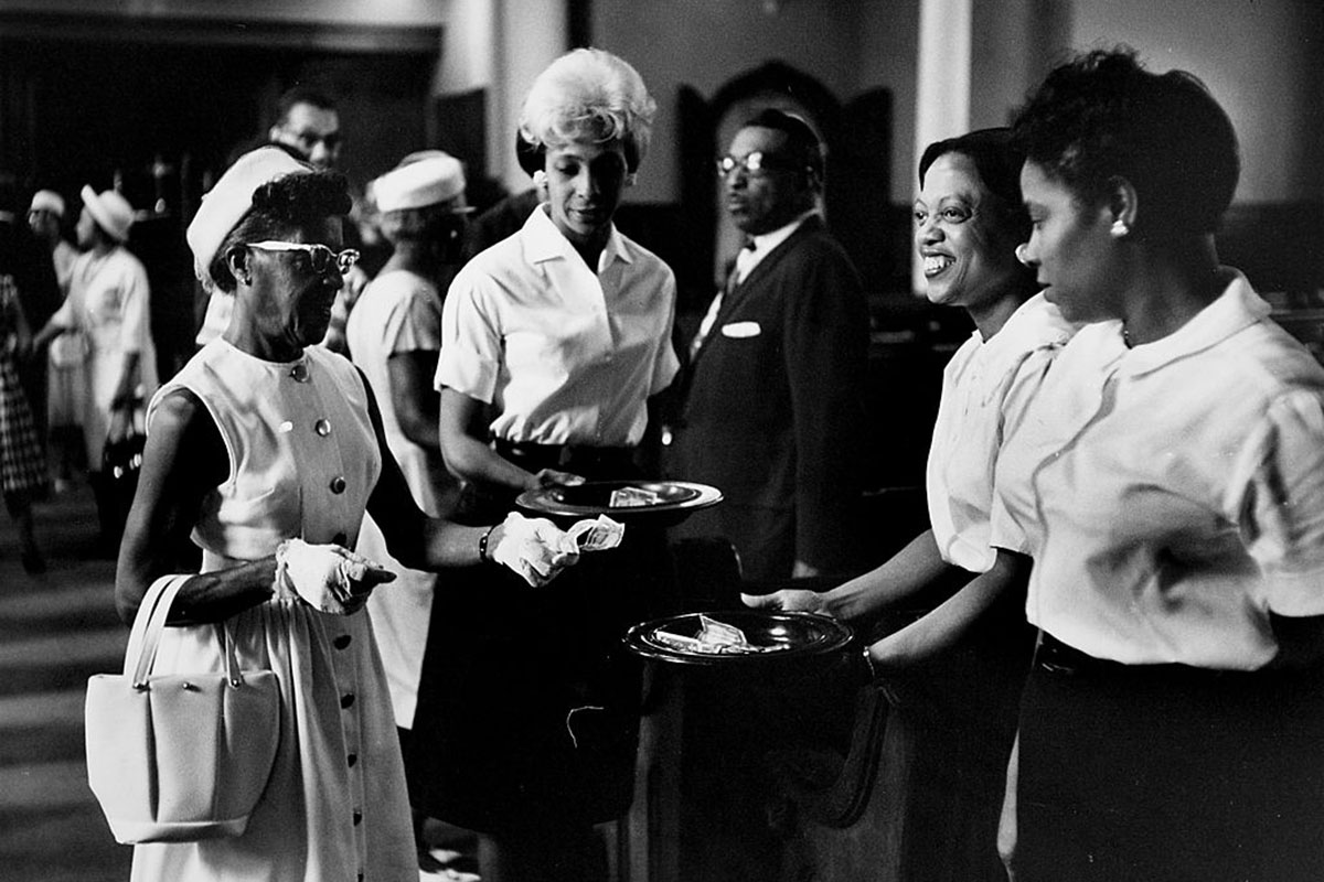A black and white photo shows several formally dressed women putting money in a church collection plate. (Black women)