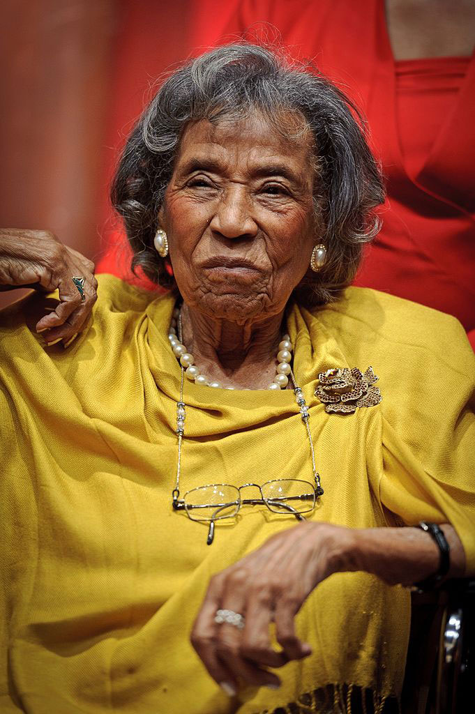 A sitting woman with gray hair in a gold-colored dress and jewelry. (Black women)