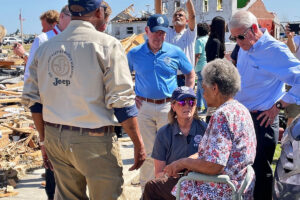 Officials speak to a seated older woman in front of destroyed houses