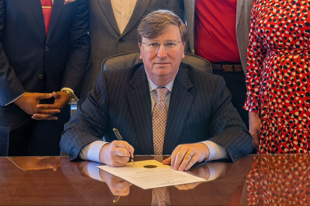 Gov. Tate Reeves seated at a table signing a piece of paper with figures behind him