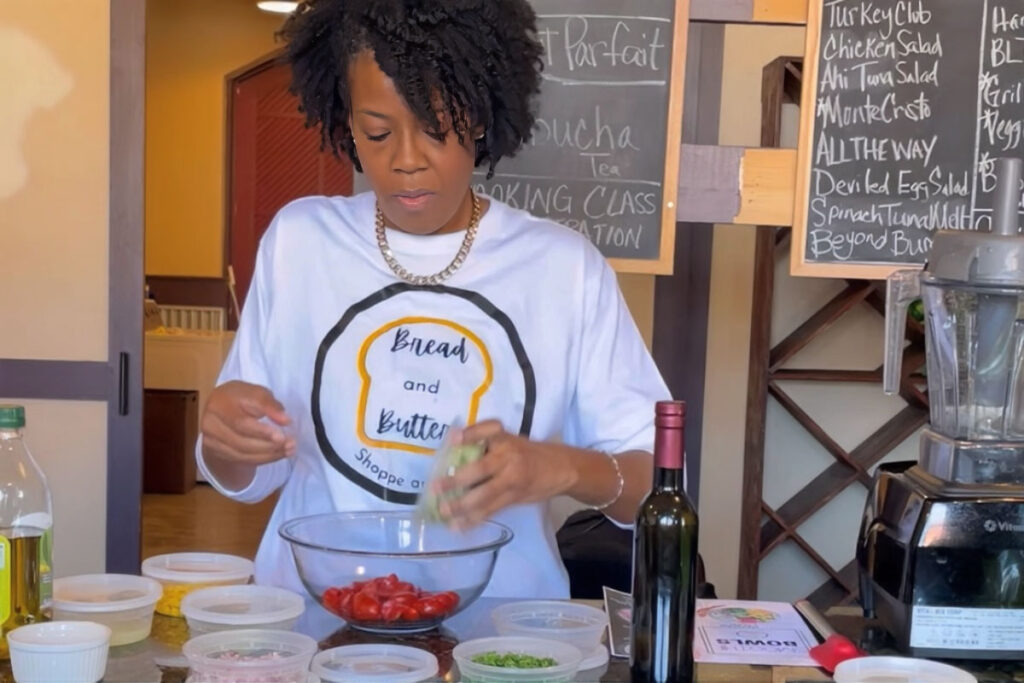A woman in tshirt that says Bread and Butter is at a counter mixing fresh ingredients in a bowl