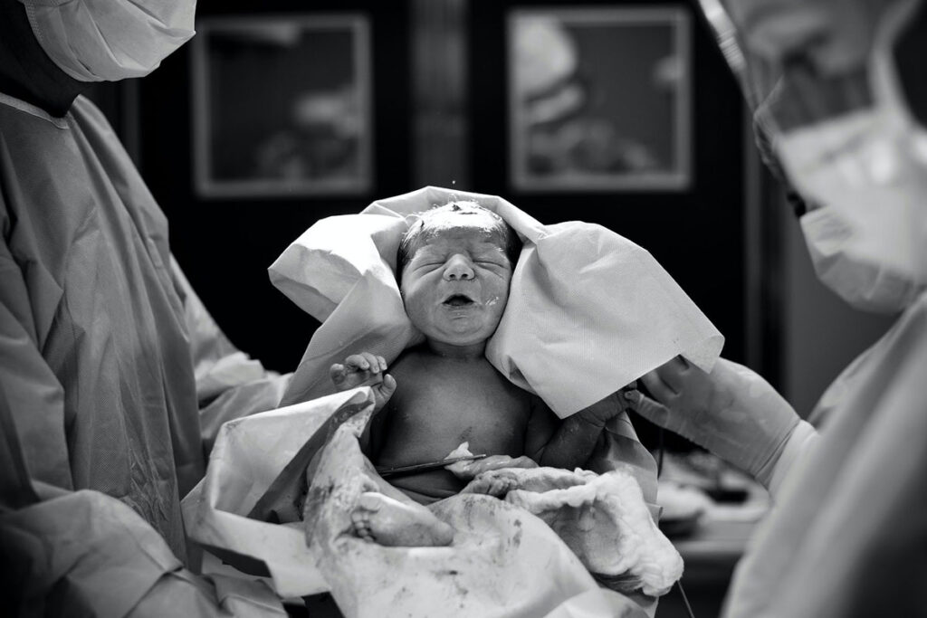 A newborn baby being held up by two medical professionals in scrub gear inside of a hospital room