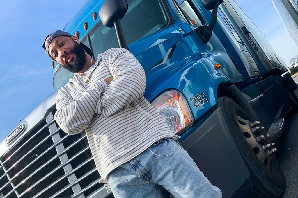 a photo of Duane Lake, a Black man, standing in front of an 18 wheeler with his arms crossed and smiling at the camera