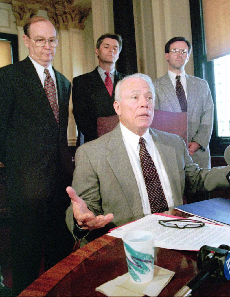 a photo of Kirk Fordice sitting at a desk with three other white men behind him