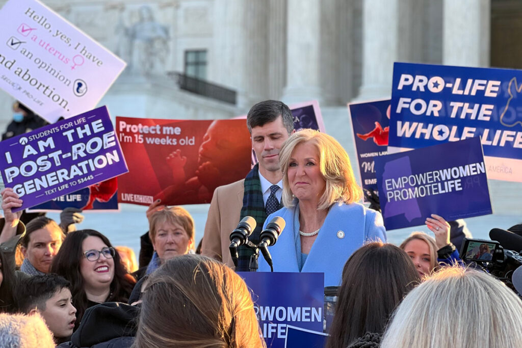 a photo of Lynn Fitch outside the U.S. Spupreme Court with anti abortion protesters holding signs up around her saying "Empower women promote life"