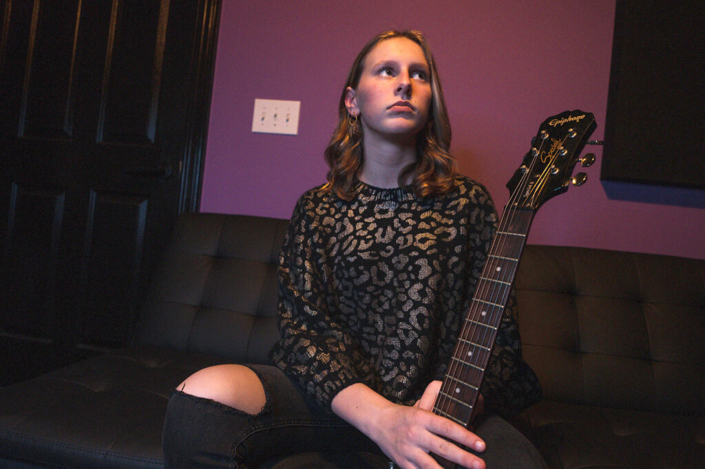 Scarlett Sullivan seated on a couch while holding a guitar, looking off to the right