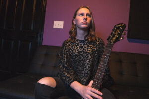 Scarlett Sullivan seated on a couch while holding a guitar, looking off to the right