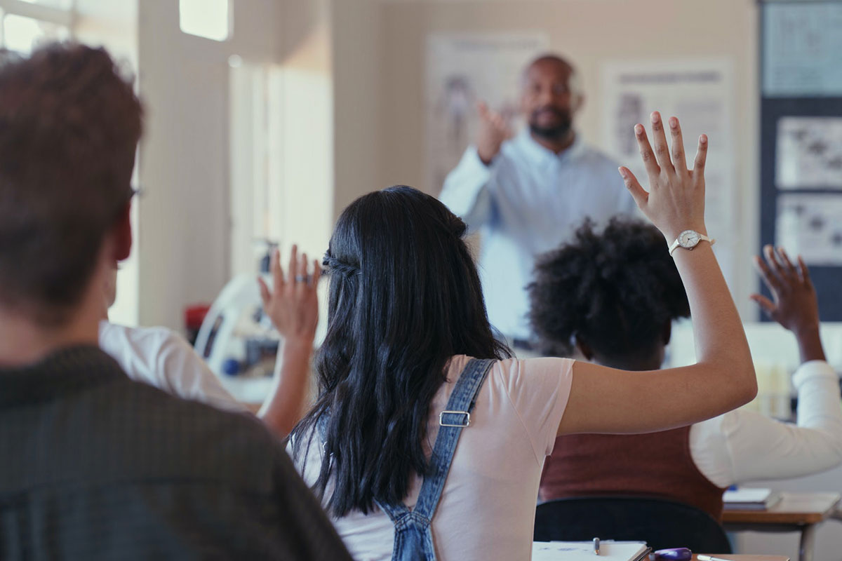 A black male teacher points to older students raising their hands in class