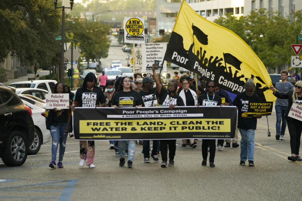 a photo of people marching in Jackson holding a "Poor People's Campaign" banner, a banner that says "Free the Land, Clean the Water & Keep It Public" and several holding signs saying "Polluted Drinking Water is Violence"