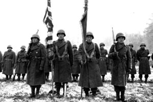 Japanese American soldiers stand at attention while holding rifles and flags