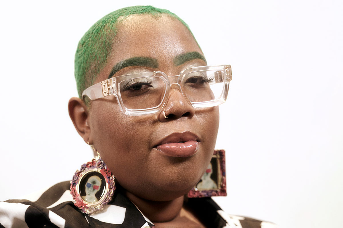 Jasmine Williams headshot, seen with green hair and clear glasses