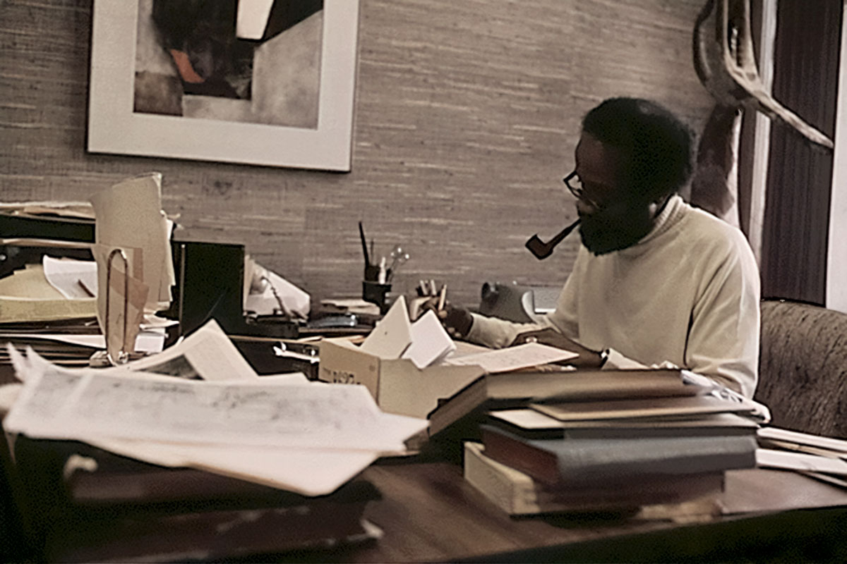 Author Lerone Bennett sitting at his desk, smoking a pipe while writing