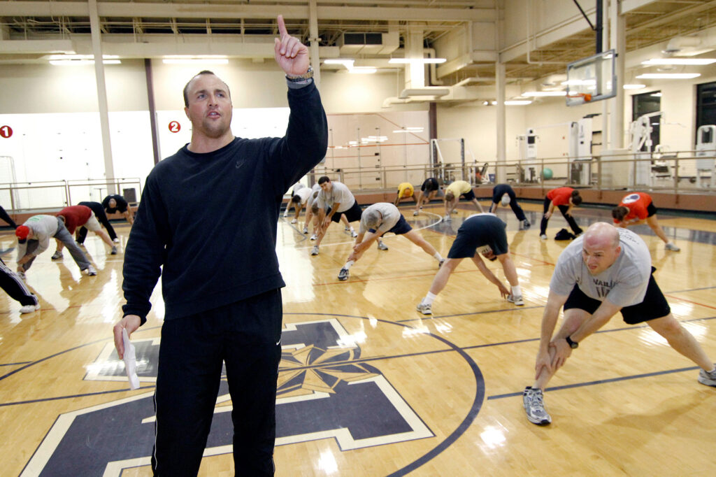 Paul Lacoste standing in a black sweatsuit as men stretch behind him in a gym