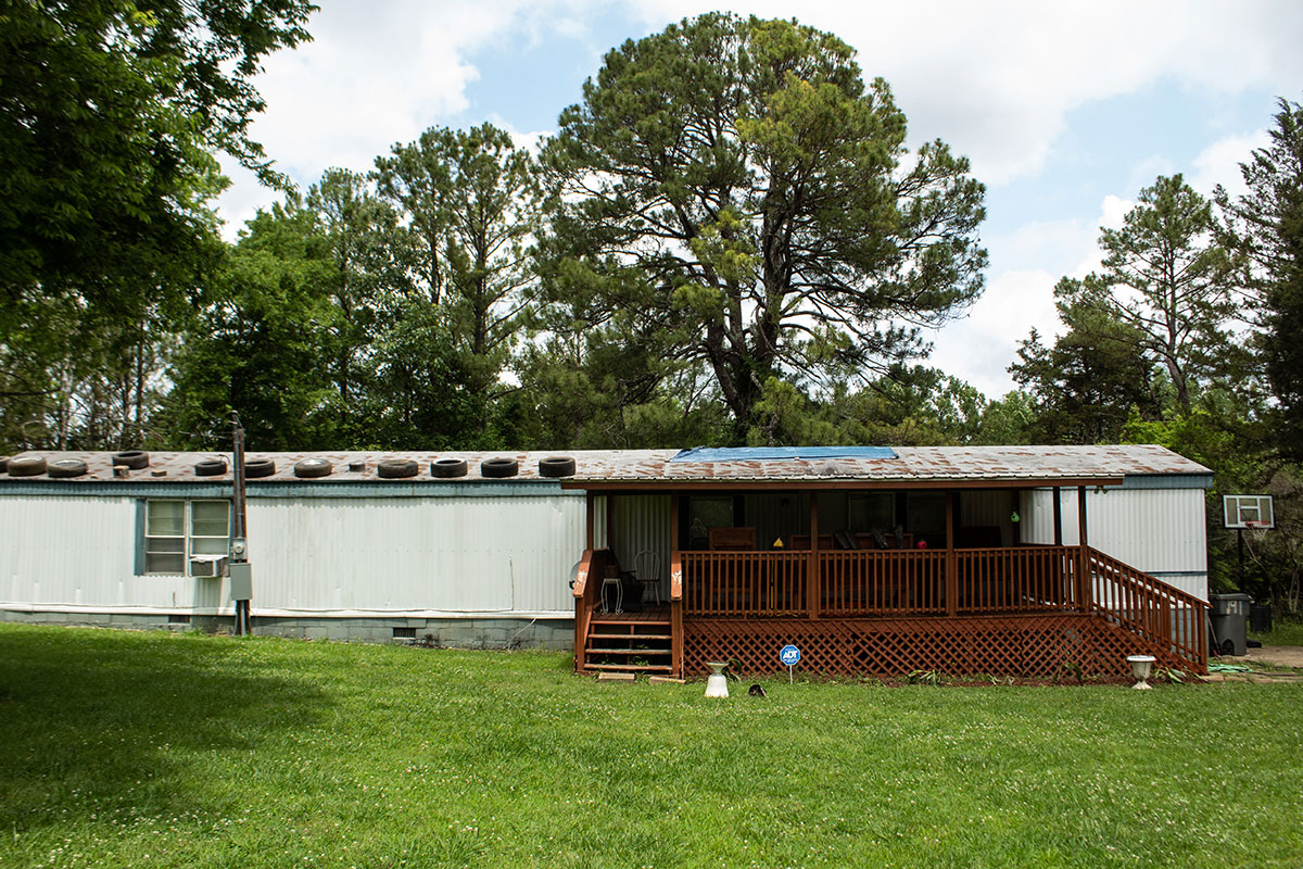 A white mobile home with a brown wooden deck attached in front, and black car tires lined on the roof