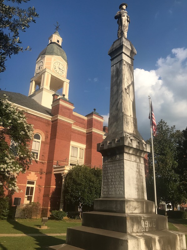 a photo of a tall Confederate monument standing in front of a courthouse, with a granite Confederate soldier standing atop it