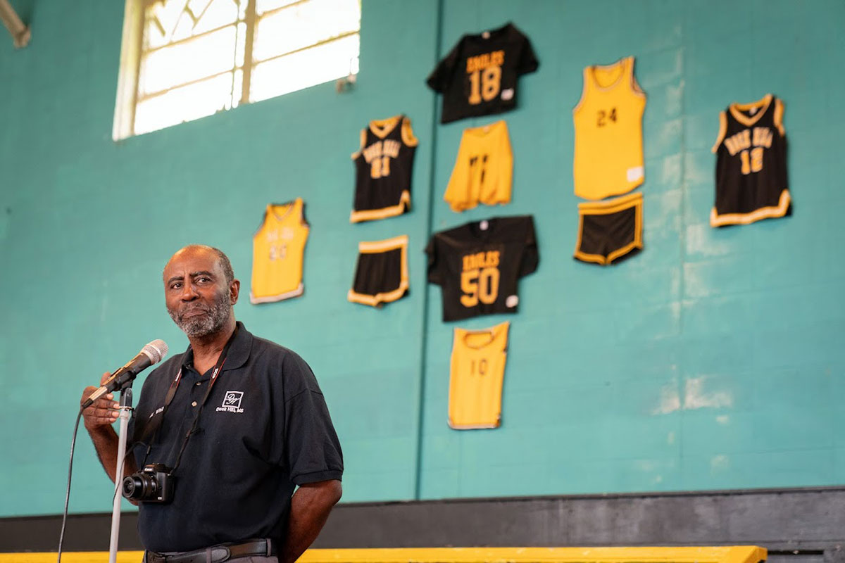 A man at a mic stands before a teal wall with black and yellow sports wear pinned to the wall