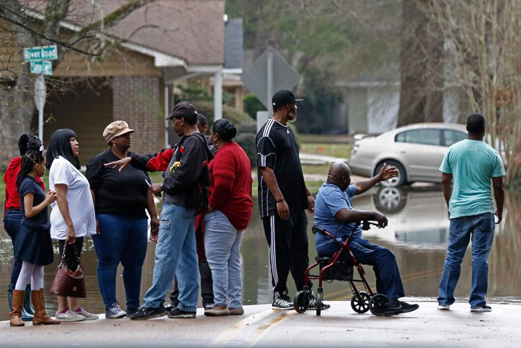 A row of people stand at the end of floodwaters in a neighborhood road