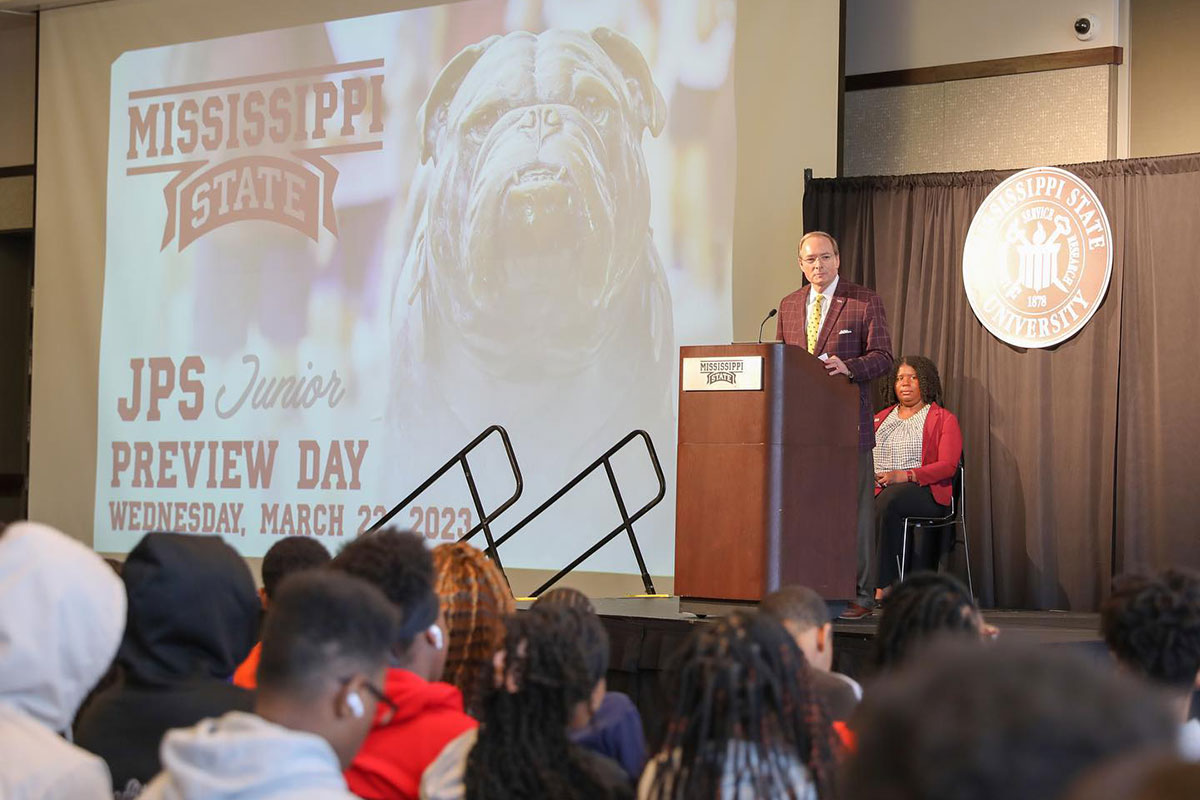 A man stands on a stage as teenagers look on from the crowd. The screen to his left says Mississippi State JPS Junior Preview Day 2023. (Black MSU students)