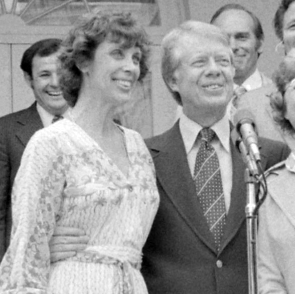 Patt Derian and Jimmy Carter stand outside in a group near a mic