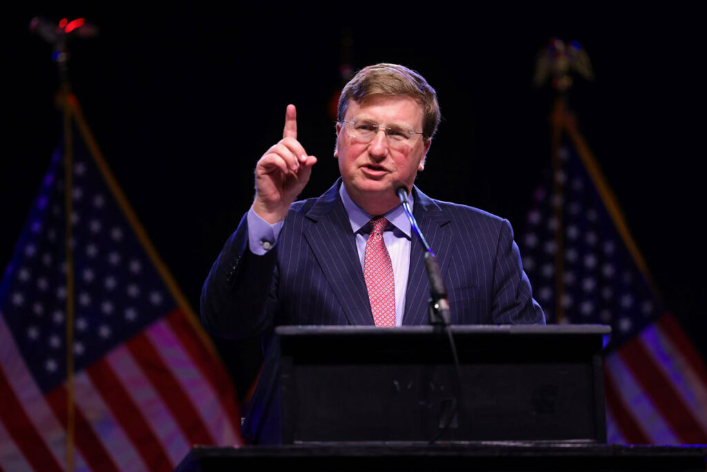 a photo of Tate Reeves at a podium in front of two American flags, holding one index finger up