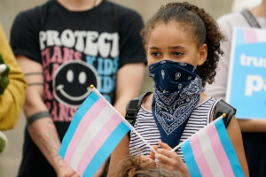 A young girl with her face partially covered holds two trans flags