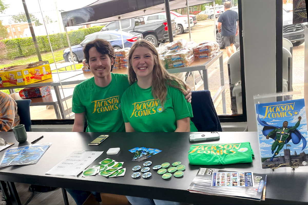 Two people in green shirts that read "Jackson Comics" sit at a table