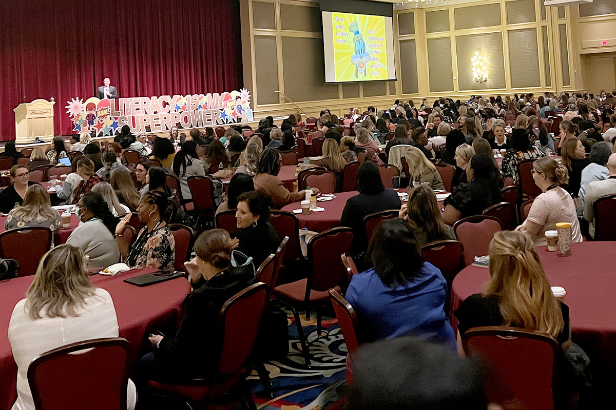 A full conference with people seated at red-covered tables. A sign reads Literacy Superpower on the stage.