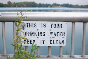 A sign in front of a lake that reads: This is your drinking water keep it clean (water challenges)