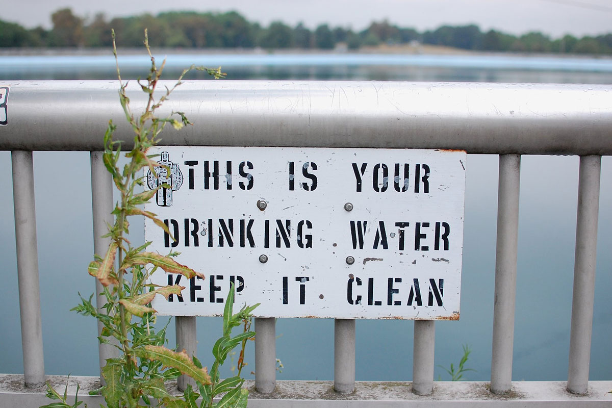 A sign in front of a lake that reads: This is your drinking water keep it clean (water challenges)