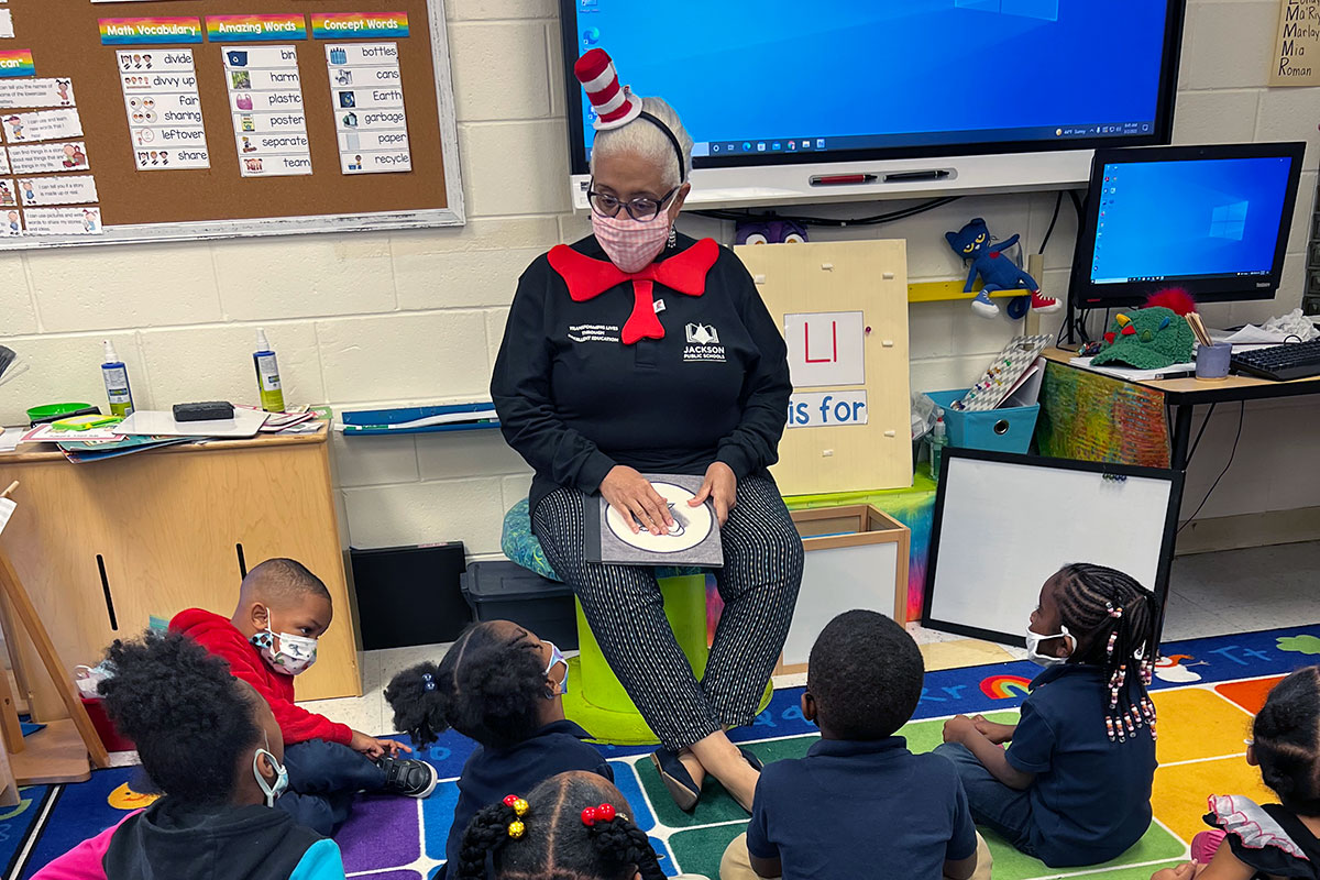 A teacher dressed like the Hat in the Hat sits before a class of children seated on a colorful floor