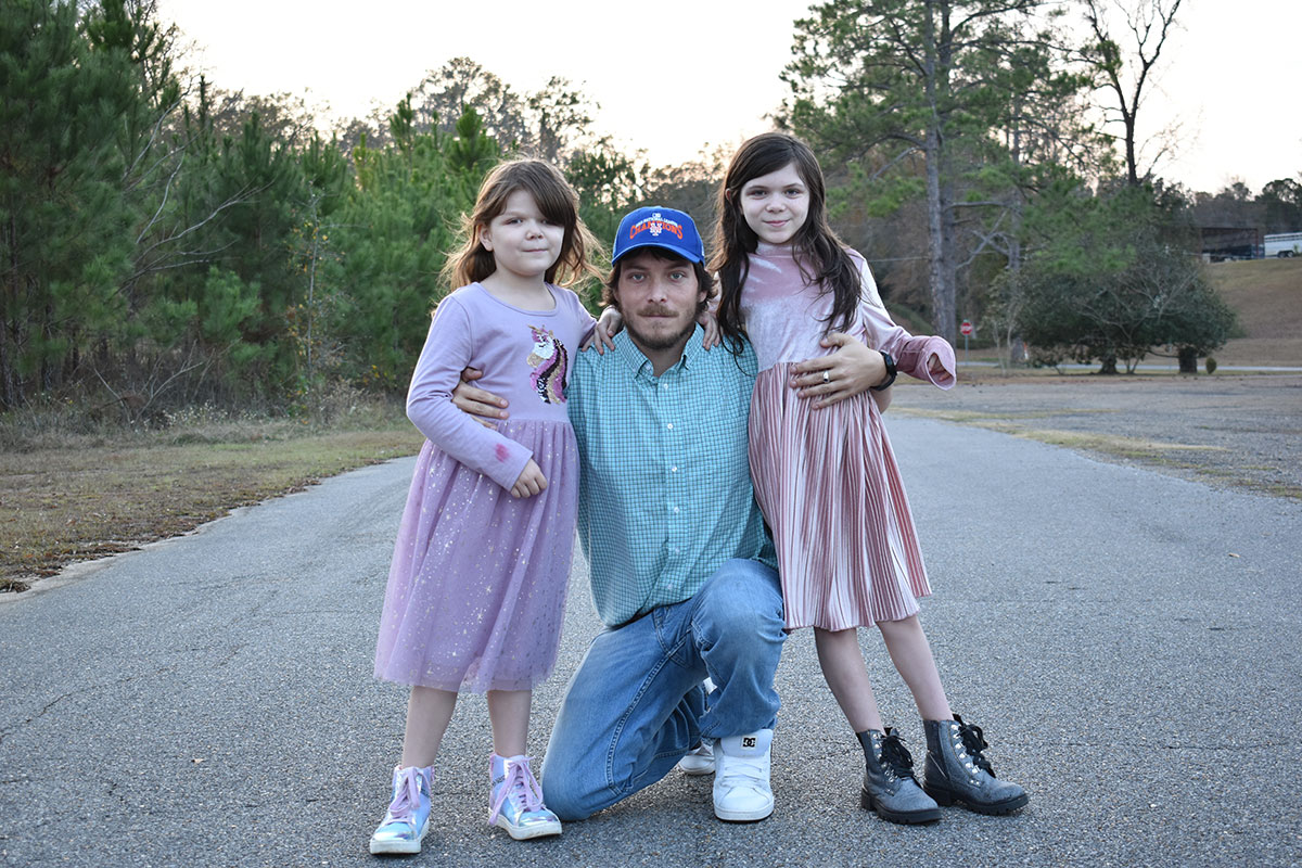 A man crouches on the ground and hugs two young girls on either side of him