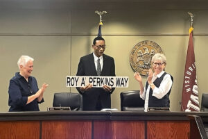 A man holding a sign that reads Roy A'. Perkins Way
