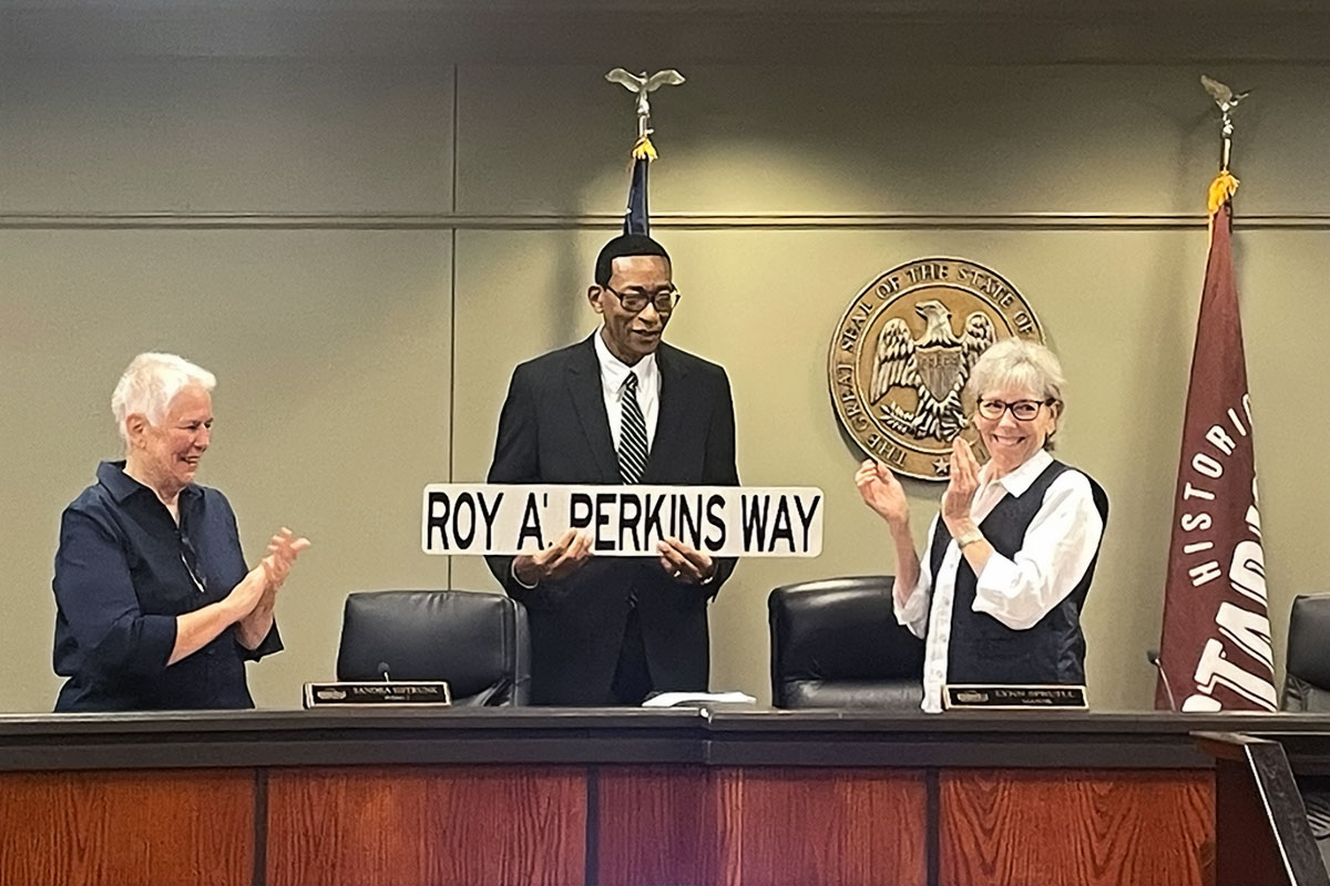 A man holding a sign that reads Roy A'. Perkins Way