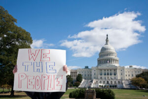 A person stands in front of the capitol with a sign that reads WE THE PEOPLE