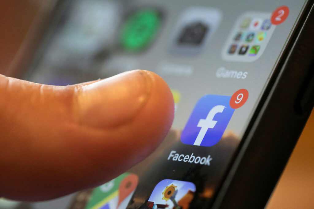 A thumb is seen about to press on the Facebook icon on a phone screen