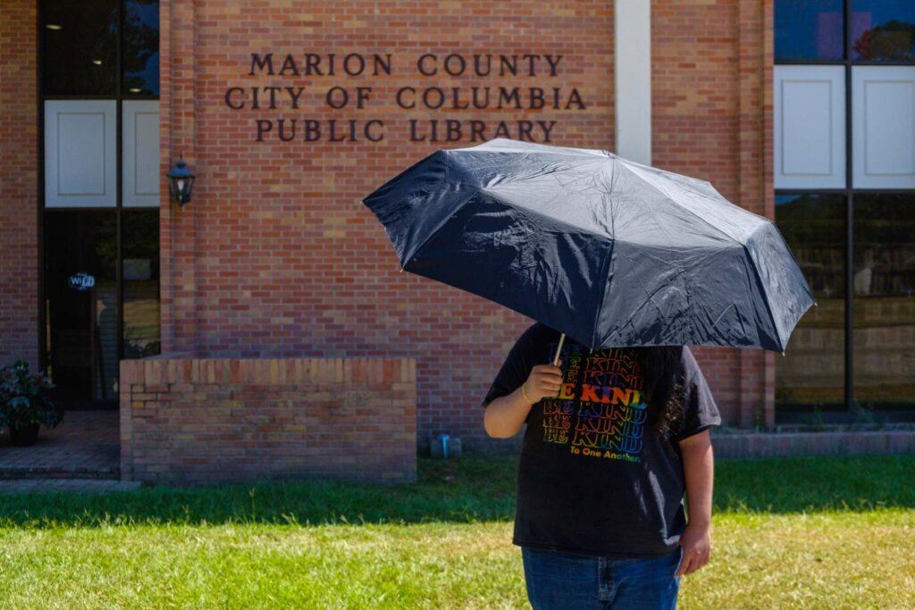 a person stands on the grass in front of a brick building that says "Marion County City of Columbia Public Library"; the person is holding a black umbrella that is covering their face, but their blue jeans and black t-shirt is visible with the words "Be Kind" in rainbow color repeated in horizontal lines down the shirt.