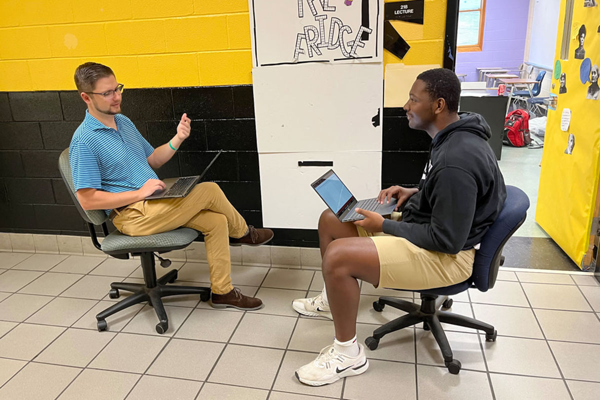 Two men sit on wheeled desk chairs in a yellow bricked school hallway