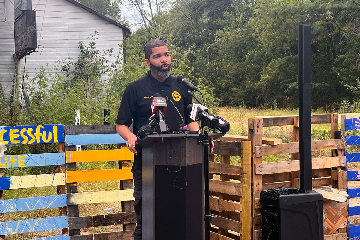 Mayor Chokwe A. Lumumba speaking at a podium outside with colorful painted fencing behind him