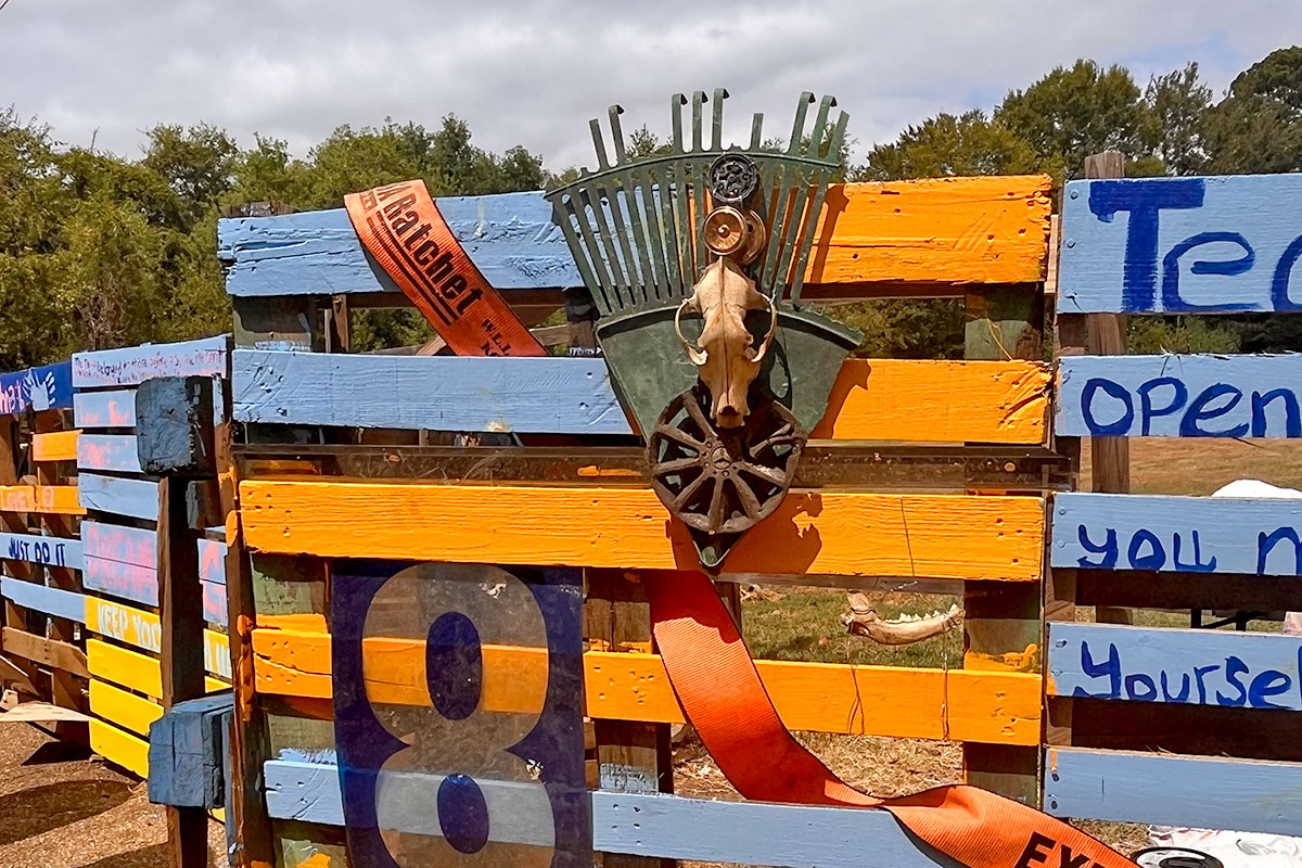 Blue and yellow painted fencing decorated with found objects like painted rakes and an animal skull