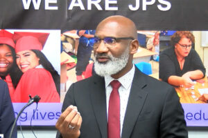 A man talks at a mic. Behind him is a banner that reads "We are JPS"