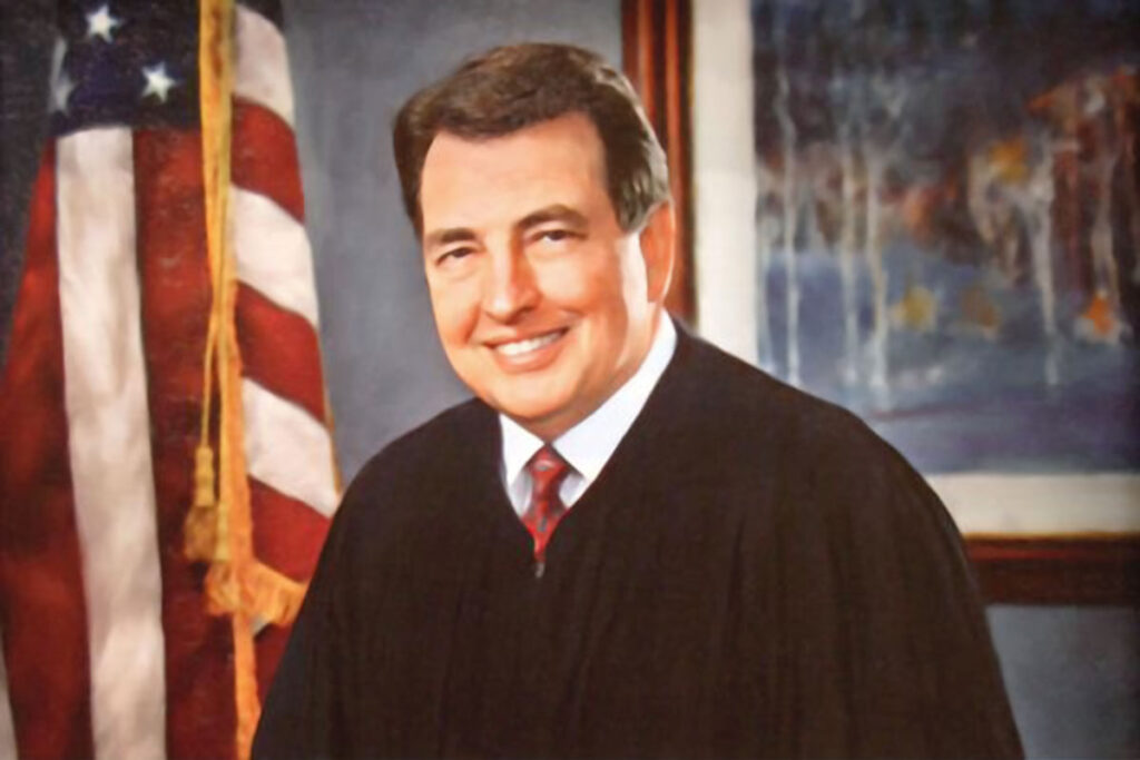 Portrait of Judge James L. Dennis inside a room with a US Flag to his left and painting to his right