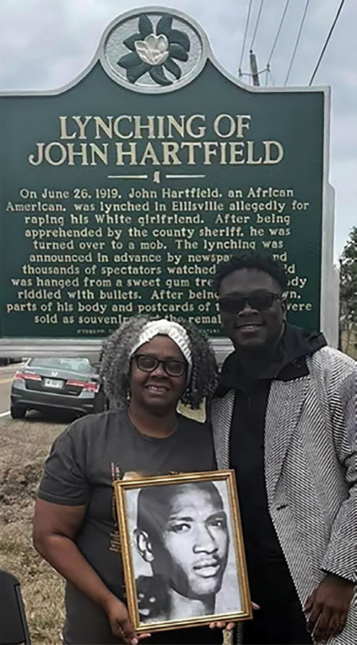 Marian Allen stands with her godson Jalen Lindsey in front of the historical marker commemorating lynching victim John Hartfield. Allen is holding a photo of Hartfield