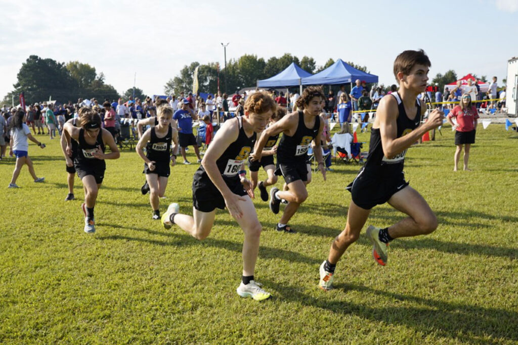 Teens in black runner uniforms run across green grass at a festival